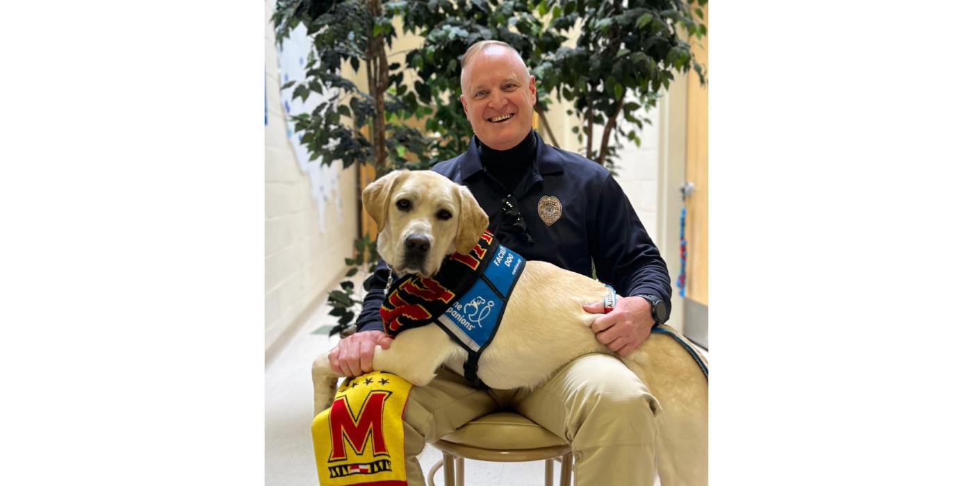 A police officer seated in a chair with his facility dog sitting across his lap.